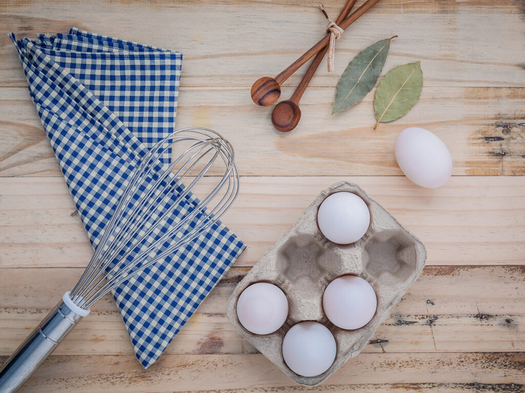 Cardboard egg box with eggs ,pepper bottle ,wooden spoons ,bay leaf and wire whisk set up on old wooden background.