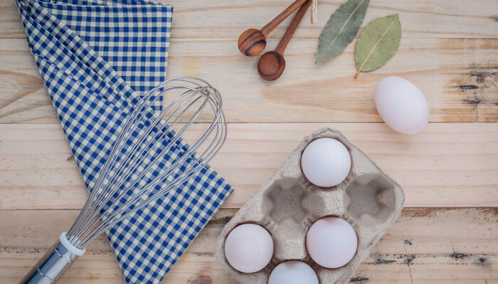 Cardboard egg box with eggs ,pepper bottle ,wooden spoons ,bay leaf and wire whisk set up on old wooden background.